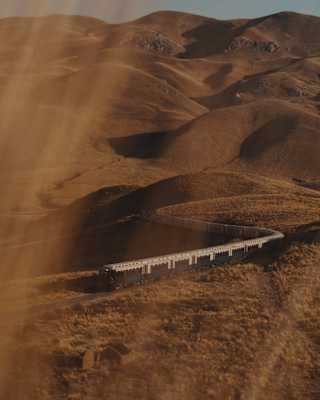 The Andean Explorer train journeys along a snaking track through undulating hills, seen from above through golden sun haze.