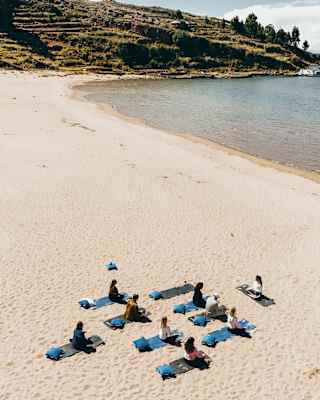 Eight guests sit cross-legged on blue exercise mats facing an instructor during a yoga session on a white-sand beach.