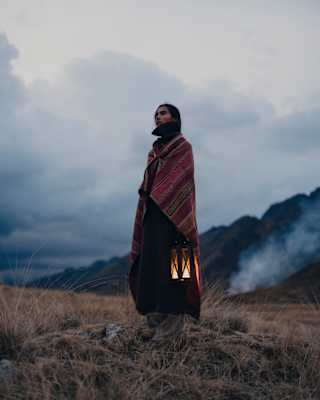 Low-angle image of a person wrapped in a woven shawl as they stand on a grassy hill in low light holding a glowing lantern.