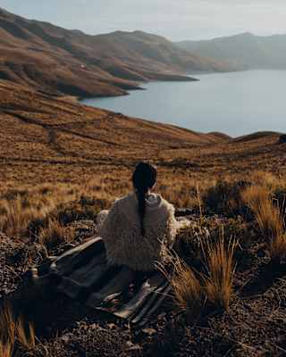 A woman with a long dark plait, wearing a wool shawl, sits on a blanket in grassland above Lake Saracocha, seen from behind.