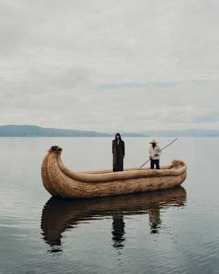 A figure in dark clothes stands in a banana-shaped vessel of bound totora reeds on Lake Titicaca, steered by a local boatman.