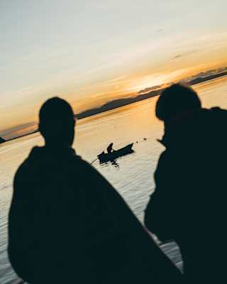 Angled image of two men gazing at a boat on Lake Titicaca, all silhouetted against the sunset-tinted waters and golden sky.
