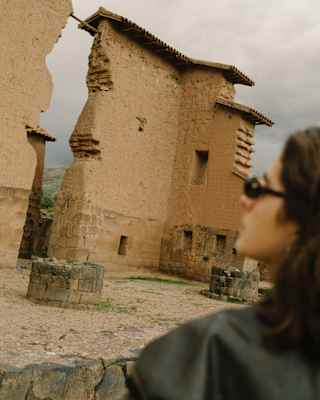 A dark-haired woman in sunglasses in soft-focus foreground gazes up at the giant walls of the Incan site of Raqchi.