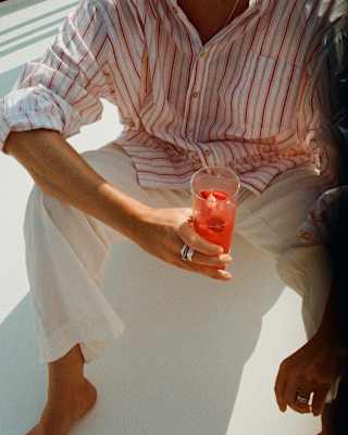 A guest, seen from the neck down in a red-striped shirt and white trousers, sits barefoot on the deck holding a pink drink.