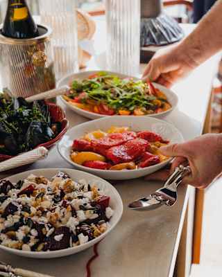 A guest reaches for bowls of roasted capsicums and bright tomato salad at a buffet table with dishes, bread and champagne.