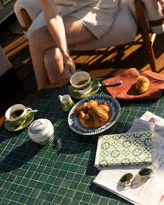 A woman sits at a low table with a book, paper, cups of tea and a bowl of croissants in an angled image of breakfast on deck.