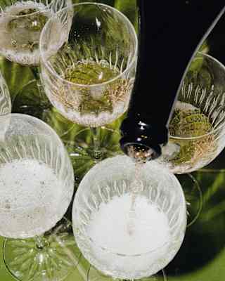 Effervescent champagne is poured evenly into six crystal glasses on the green lounge table, seen close-up from above.