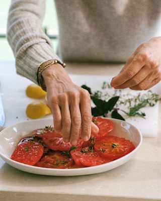 Chef Dominique Crenn prepares a salad of sliced ripe beef tomatoes sprinkled with herbs, seen in close-up.