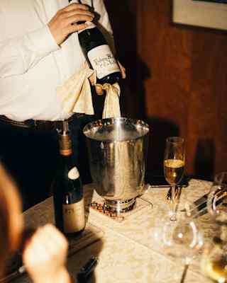 At a table laid for dining, with wineglasses and gold-plated cutlery, a waiter shows guests the label on a bottle of wine.