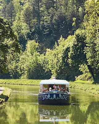The Fleur de Lys cruises up the serene Ouche river, bordered by big trees and green banks, where a cyclist enjoys the route.