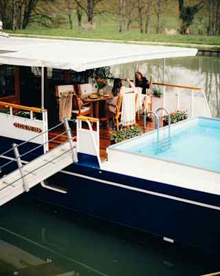 Guests relax round a table on the deck while the boat is moored, with a gangplank to the canal bank.