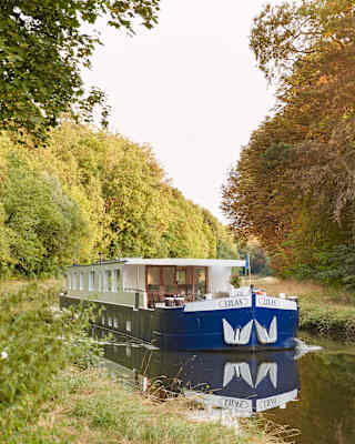 Front view of a luxurious river barge sailing down a sun-splashed tree-lined canal