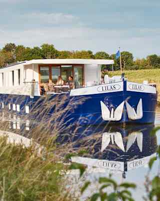 The Lilas cruising with two passengers on deck enjoying the sunshine and tranquillity, pictured through grasses on the bank.