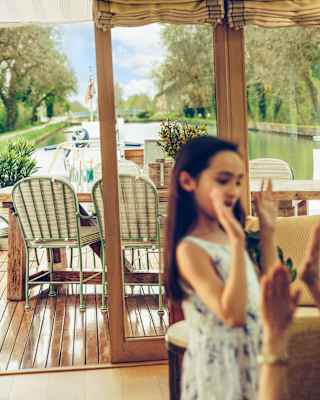 A girl plays a hand-clapping game in the living area in soft-focus, with focus on the deck behind and views of the waterway.