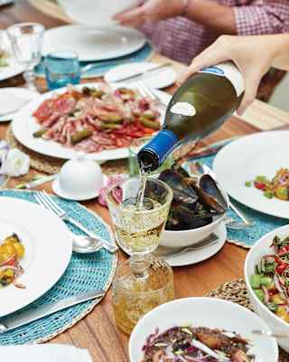 White wine is poured into a crystal glass as guests enjoy a feast, with plates of charcuterie, lentils, salad and shellfish.