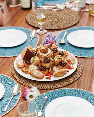 A piled plate of sugar-dusted Madeleines and choux buns with creme and chocolate rests on a laid table with blue place mats.