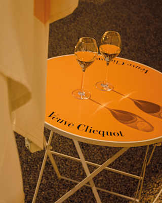 Two glasses of sparkling champagne cast their shadows on the vibrant orange top of a Veuve Cliequot-branded table.
