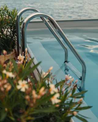 Chrome steps lead into the pool, where smooth water contrasts with the rippling waterway behind, seen over potted plants.