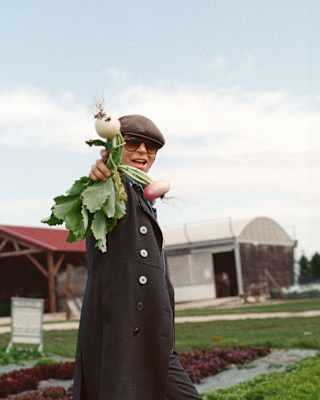 Chef Dominique Crenn, in dark glasses and a flat cap, stands in a vegetable farm, holding up a bunch of fresh-pulled turnips.