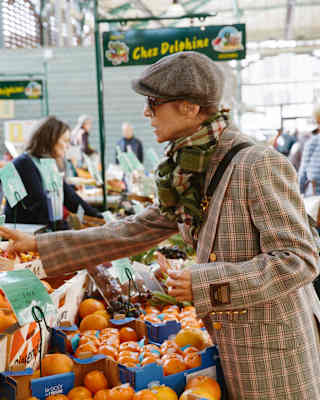Chef Dominique Crenn, in checked jacket and flat cap, leans over orange crates as she selects produce at a market stall.