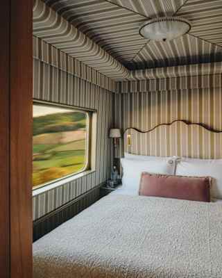 A cosy bed with white bedding and a pink accent cushion sits in a train compartment with striped wallpaper. A window shows a blurred outdoor landscape, indicating the train is moving.