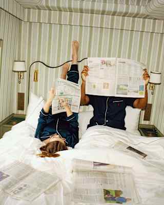 Two people in pyjamas relax on a bed in a striped train compartment, reading newspapers. One is lying with feet up against the wall, while the other sits upright. More newspapers are spread across the white bedding.