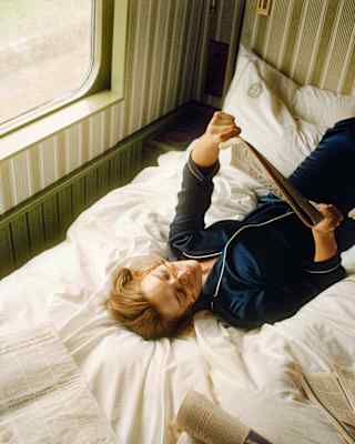 A person in pyjamas lies on a bed near a window, reading a newspaper, with more newspapers scattered around them. Natural light streams in from the window, creating a cosy and relaxed atmosphere.