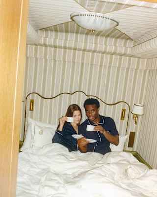 A woman and a man sit together on a bed with white sheets, leaning against the headboard, both drinking from white cups in a warmly lit, striped wallpapered room.
