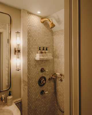 A small, elegant bathroom with patterned tiled walls, a built-in showerhead, wall-mounted soap dispensers, a round mirror, a modern basin, and a vertical light fitting beside the mirror.