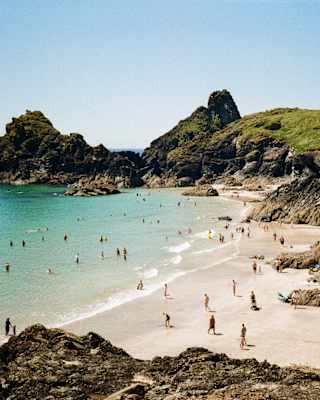 People relax and swim at a sandy beach surrounded by rocky cliffs and clear blue water under a sunny sky. Green hills rise in the background.