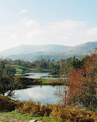 A tranquil landscape with a calm lake, trees with autumn leaves, and rolling hills in the background under a partly cloudy sky. The scene is peaceful and bathed in soft daylight.