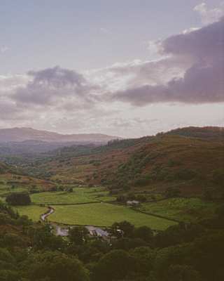 A wide, scenic landscape of rolling green hills and valleys under a cloudy sky, with distant mountains on the horizon and patches of sunlight illuminating fields and trees.