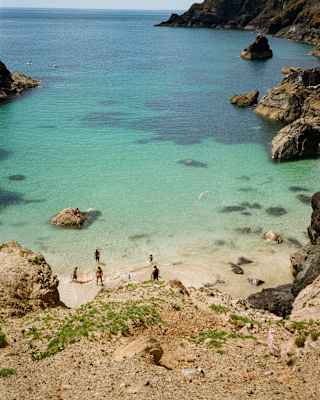 A small sandy beach with turquoise water, surrounded by rocky cliffs. A few people are standing at the shore. The sky is partly cloudy and the scene is bright and peaceful.