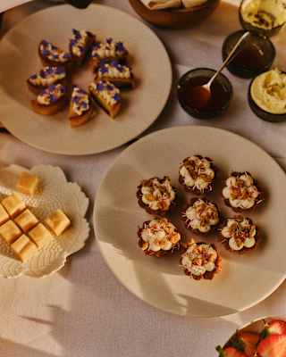A table set with assorted desserts, including mini tarts topped with cream and fruit, small squares of fudge, pastries with purple flowers, strawberries, and metal tongs on a white tablecloth.