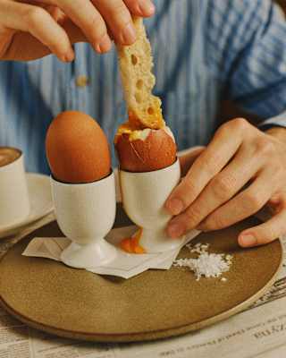 A person in a blue striped shirt dips a piece of toast into a soft-boiled egg in an egg cup, with another egg, salt, and a brown plate on a table covered with newspaper.