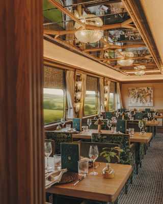 Elegant train dining car with wooden tables set for meals, large windows with scenic countryside views, green upholstered chairs, and mirrored ceiling with ornate lighting.