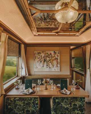 Elegant dining area inside a vintage train carriage with ornate lighting, floral-upholstered chairs, laid table, large windows with passing countryside views, and a mirrored ceiling reflecting warm interior lighting.
