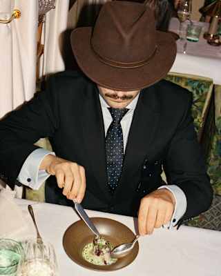 A person in a suit, tie, and brown hat sits at a restaurant table, looking down whilst eating from a bowl with a spoon and fork. The setting appears formal, with elegant tableware and décor.