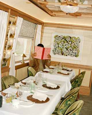 A person sits alone at a long dining table set for several people, holding up a red menu and obscuring their face. The room is elegantly decorated with green chairs, white tablecloths, and floral wall art.
