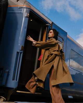 A woman in a brown coat and wide-legged trousers is stepping onto a blue train, with a partly cloudy sky in the background. She is looking up and reaching towards the train door.