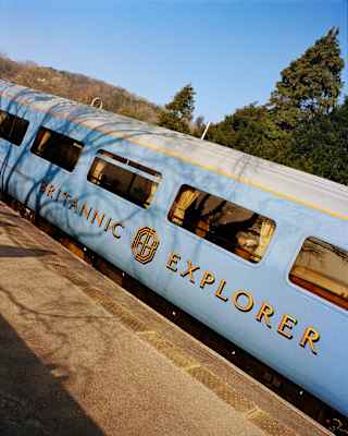 A blue railway carriage labelled BRITANNIC EXPLORER is stopped at a station platform on a clear day, with trees and a hill visible in the background.