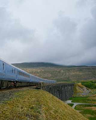 A blue passenger train travels across a bridge in a rural, hilly landscape with cloudy skies overhead.