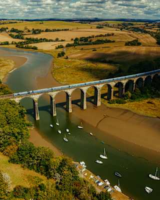 An aerial view of a train crossing a stone railway viaduct over a river with small boats moored in the water, surrounded by rolling hills and fields.