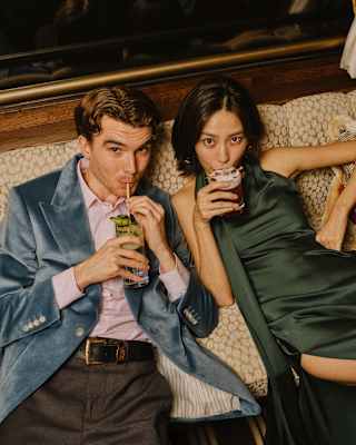 A man in a blue velvet blazer and a woman in a green dress sit closely on a sofa, both sipping drinks and looking up towards the camera with relaxed expressions.