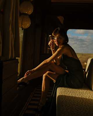 A woman in a dark dress sits on the edge of a seat by a window, bathed in warm sunlight, with her legs crossed and her hand resting on her face, inside a vintage-style room or railway carriage.