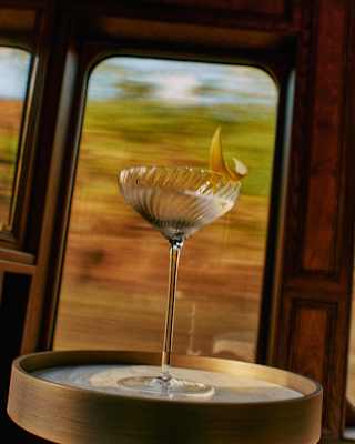 A crystal cocktail glass with a lemon twist sits on a small round table inside a moving train, with blurred scenery visible through the window in the background.