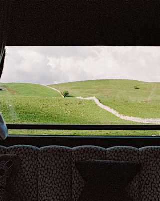 A green hillside with a rocky path is seen through a large window framed by dark curtains, behind a patterned sofa with cushions inside a cosy room.