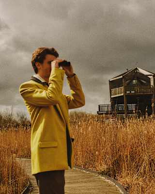 A person in a yellow coat uses binoculars on a wooden path through tall grass, with a wooden observation tower and dark, cloudy sky in the background.