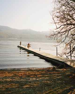 A wooden jetty extends into a calm lake, with two people standing near the end. Ripples spread across the water, and tree branches frame the right side. Distant hills are visible under a hazy sky.