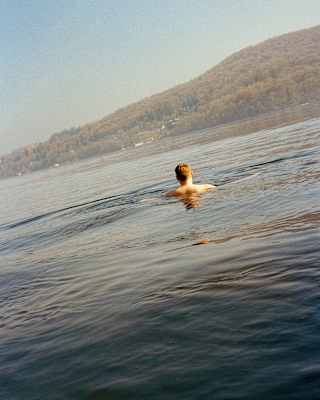 A person swims alone in a calm lake, facing away from the camera, with a tree-covered hill in the background under a clear sky.